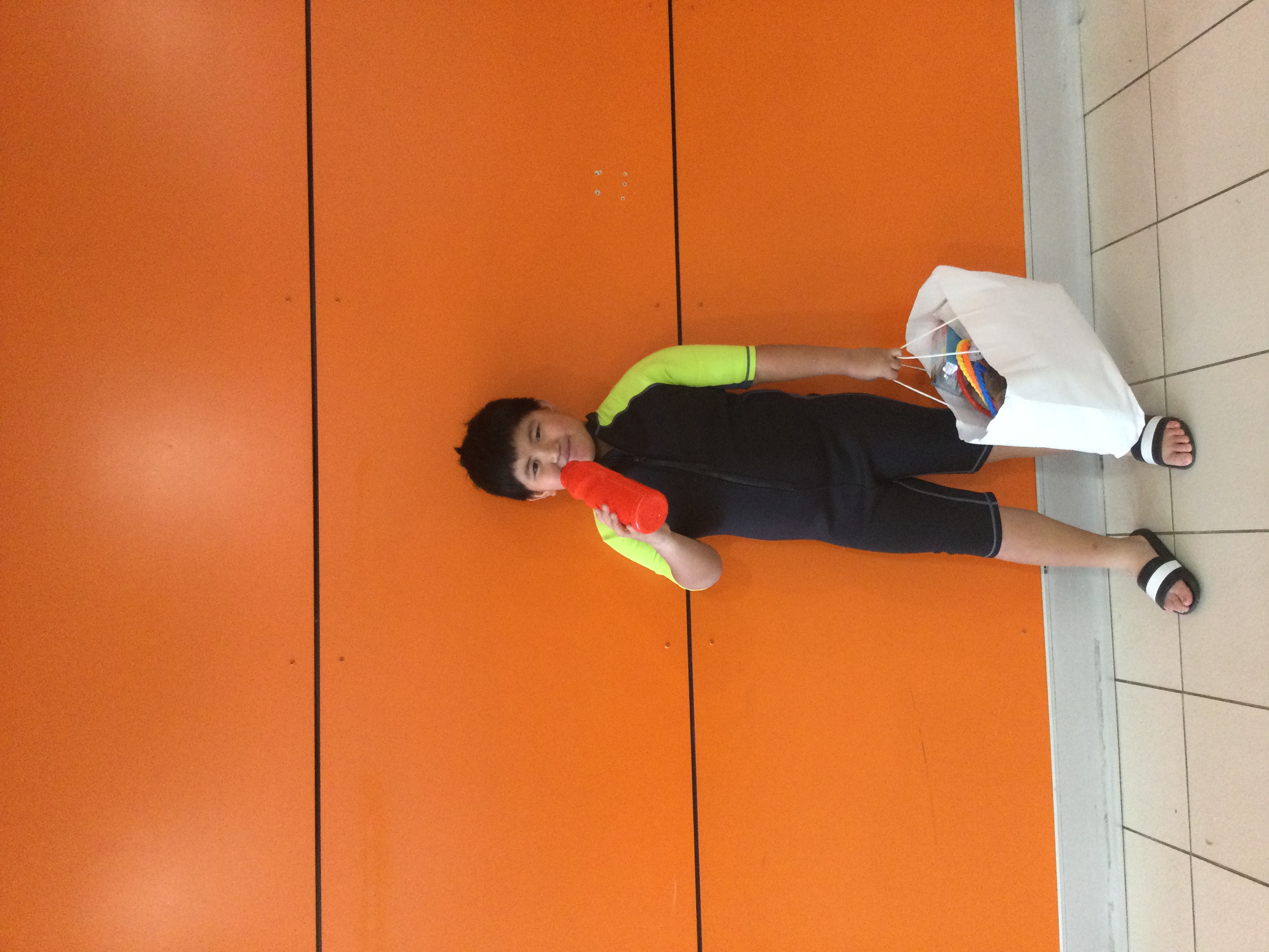 Young boy with black hair in a black swimsuit standing in front of an orange wall holding a white bag with his left hand and holding a red water bottle to his mouth with the other