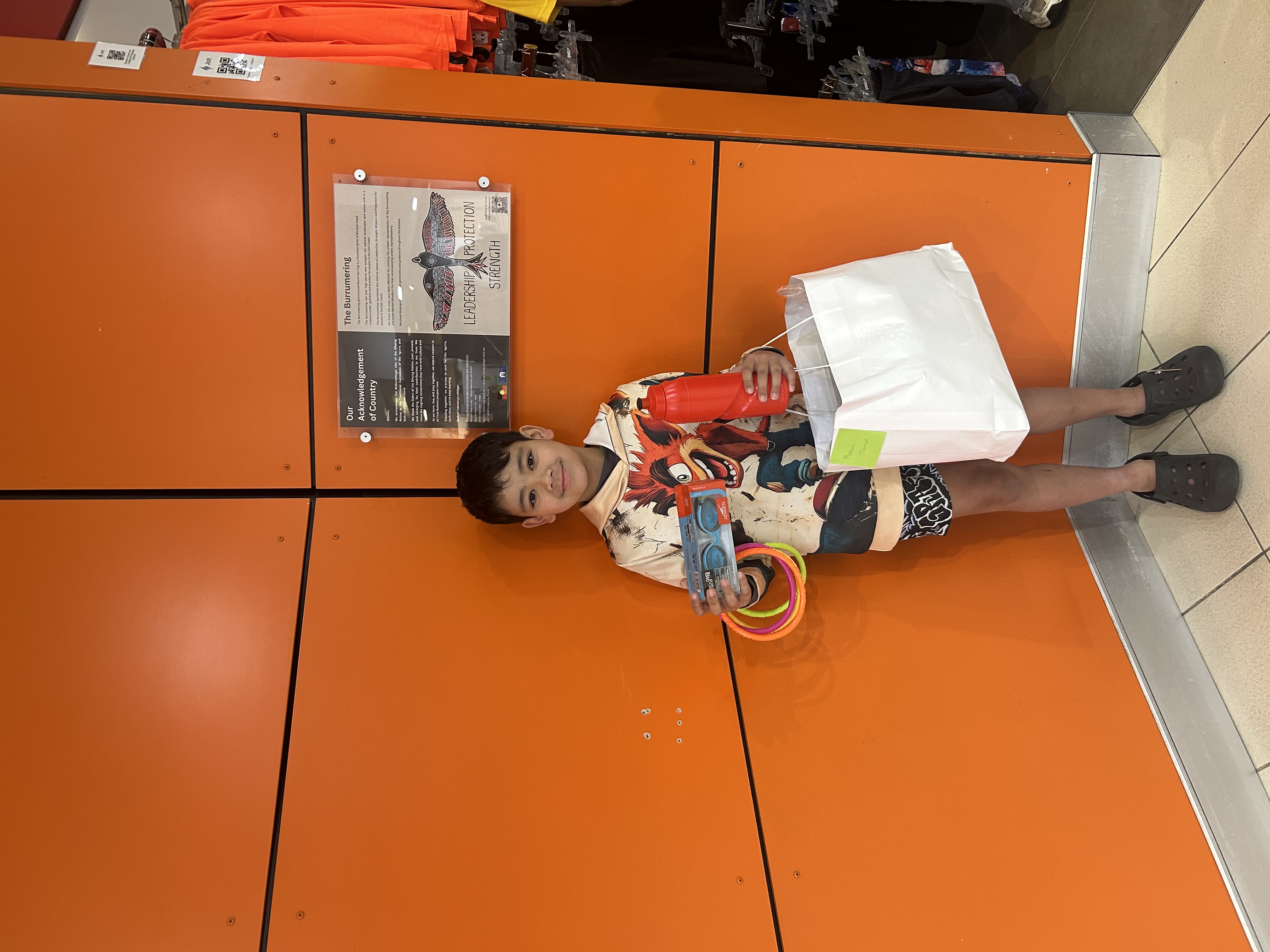 A young boy with dark hair standing in front of an orange wall with his arms full of swimming gear including goggles, diving rings and a water bottle