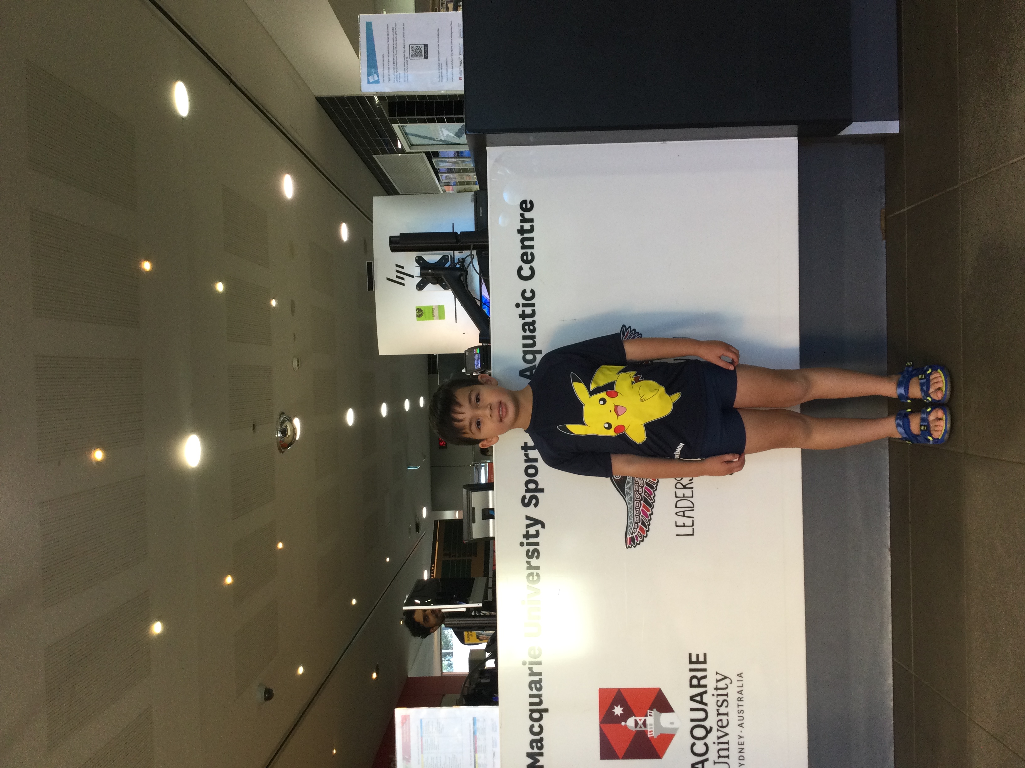 A young boy with dark hair wearing a shirt with a yellow animal in the centre of the shirt and black shorts standing it front of a white and grey desk