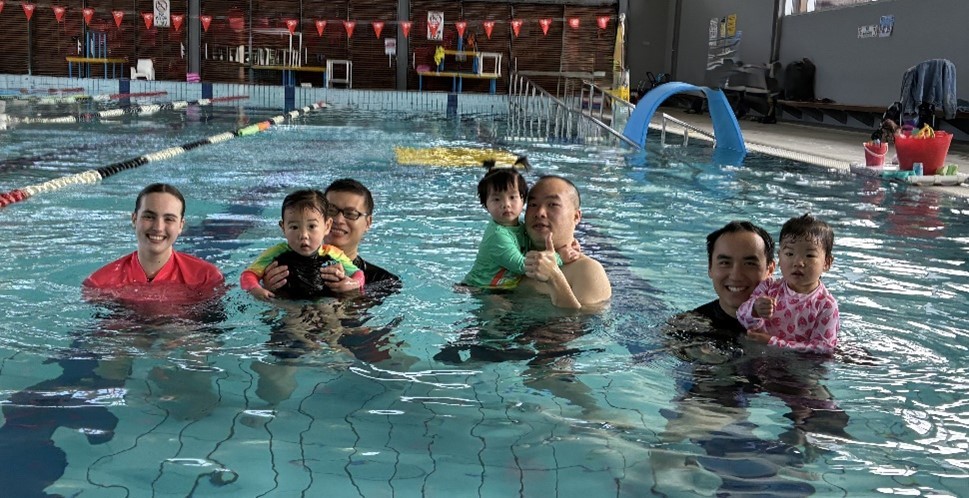 3 sets of parents in an indoor pool holding their young children alongside a Learn to swim instructor in a bright pink rashie 